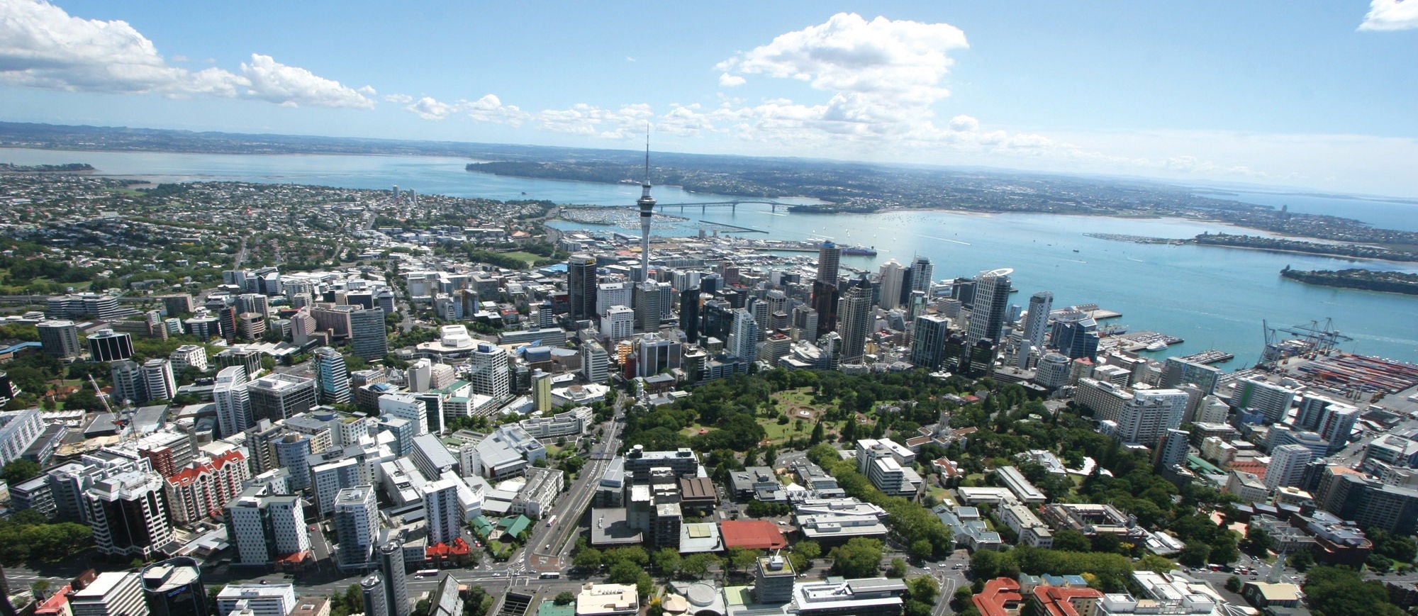 An elevated view looking north west over the Auckland CBD. It is a blue sky day with a few puffy clouds, and the skyscrapers of Auckland Tāmaki Makaurau shine in the morning sun, with the harbour bridge and north shore in the back ground.